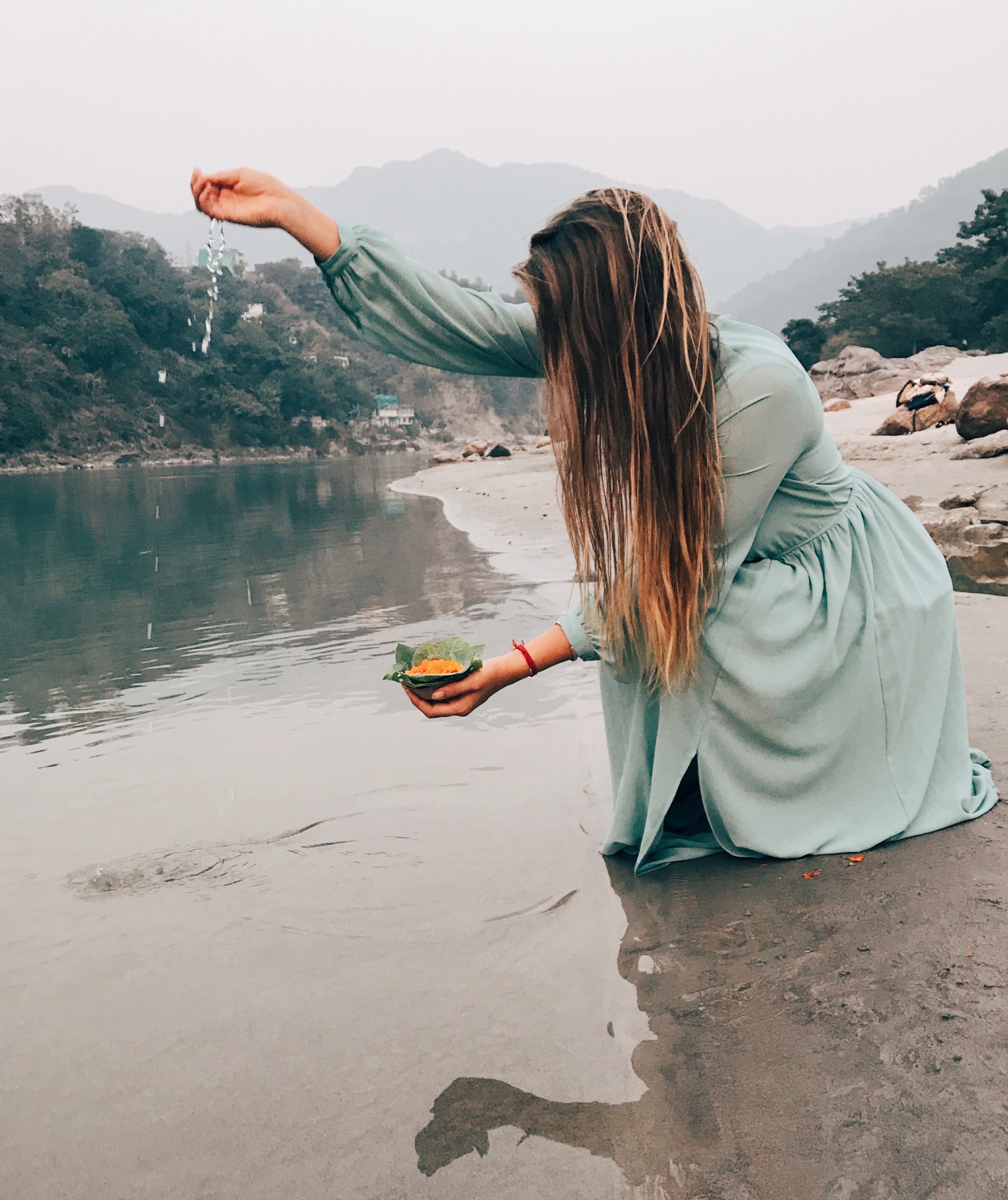 Erika kneeling at the edge of a river in a long green dress, holding a leaf bowl of marigold petals as water drips from her hand