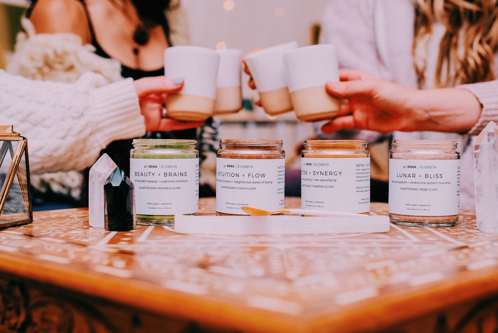 Four jars of Erika's adaptogenic elixirs on a carved wooden table while friends toast with ceramic cups
