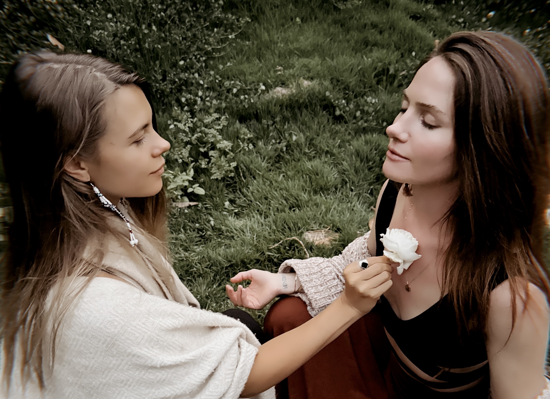 Two women sitting in a grassy field, one holding a white flower to the other's heart