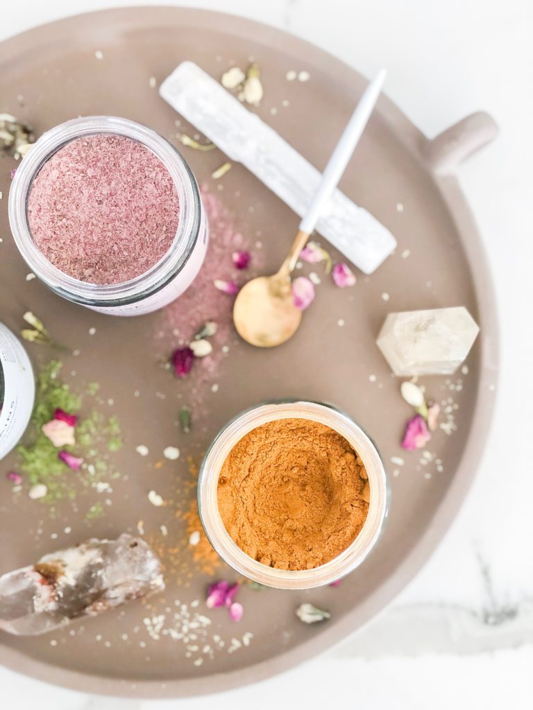 Overhead flat lay of open elixir jars with rose and golden powders on a round tray with crystals and dried petals