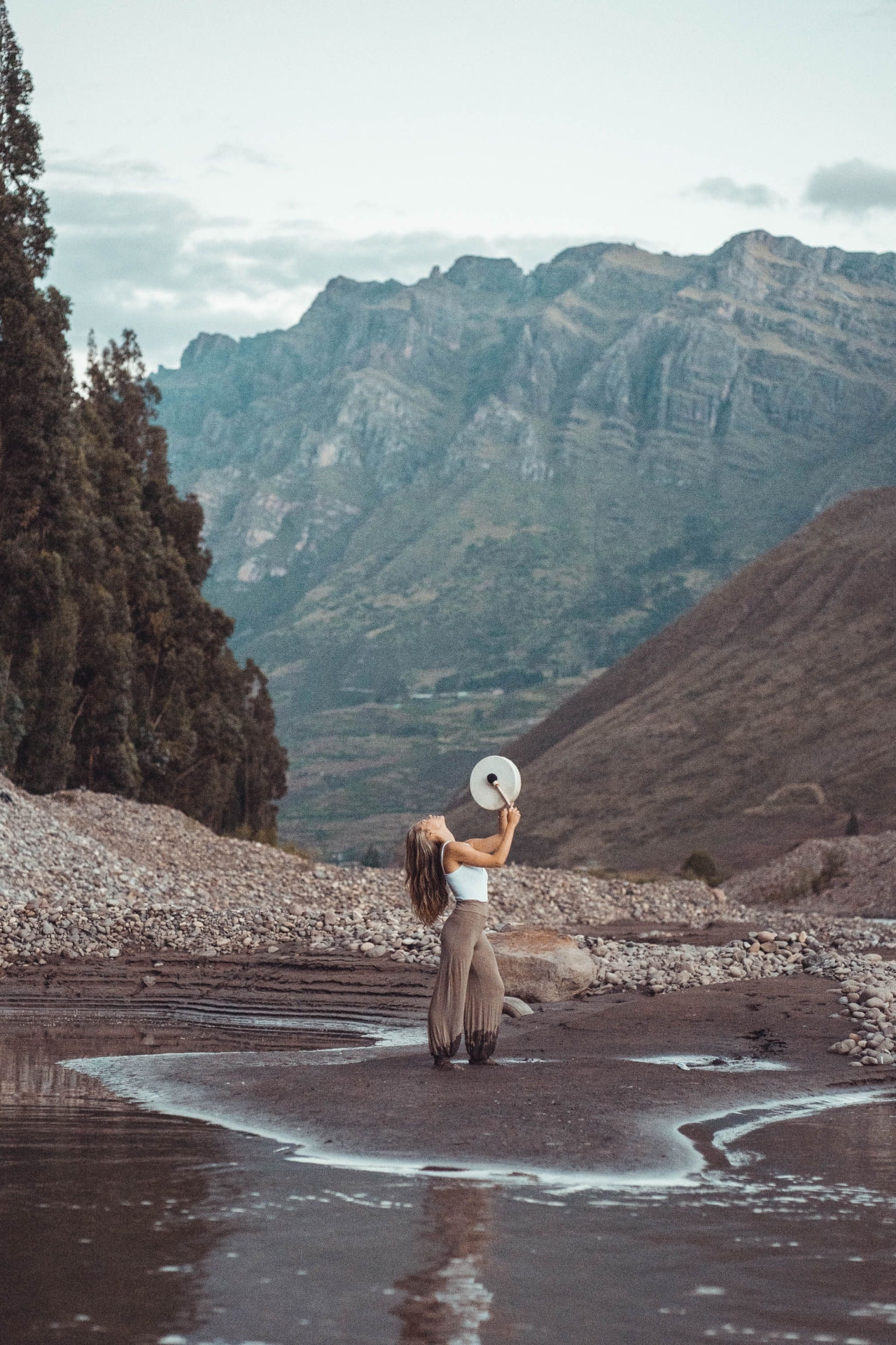 Erika standing barefoot on a mountain riverbank, lifting a white frame drum toward the sky with peaks rising behind her