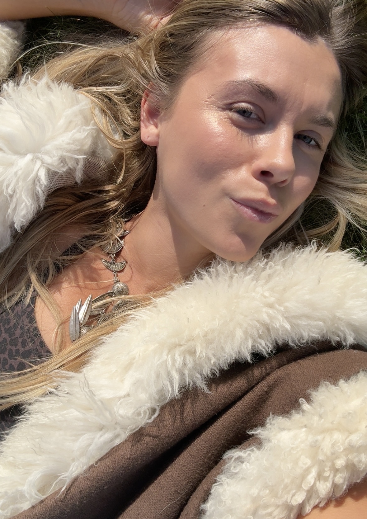 Erika lying in the sun on grass, wearing a brown coat trimmed with cream shearling and a silver feather necklace