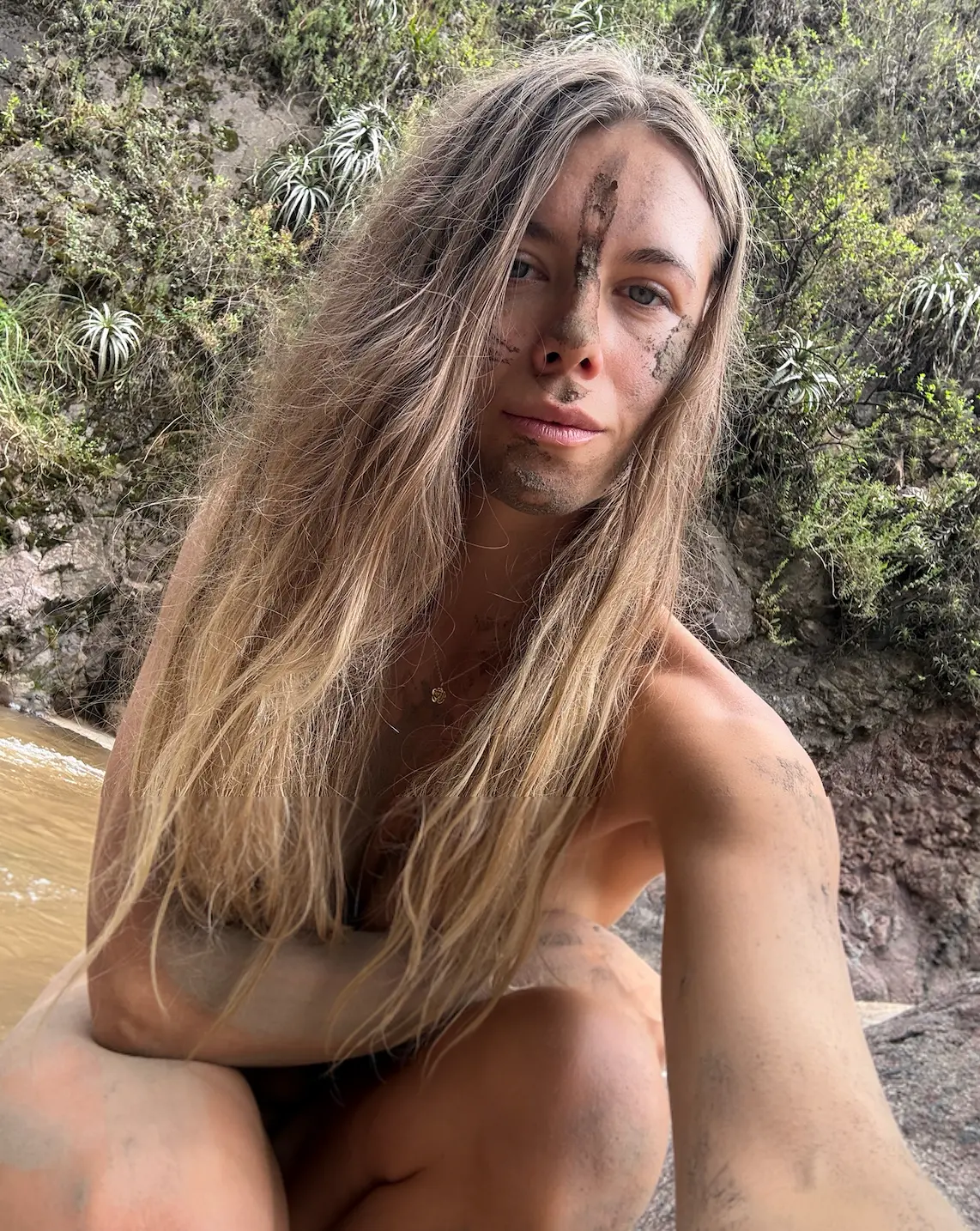 Erika with mud on her face, sitting by a river in the jungle