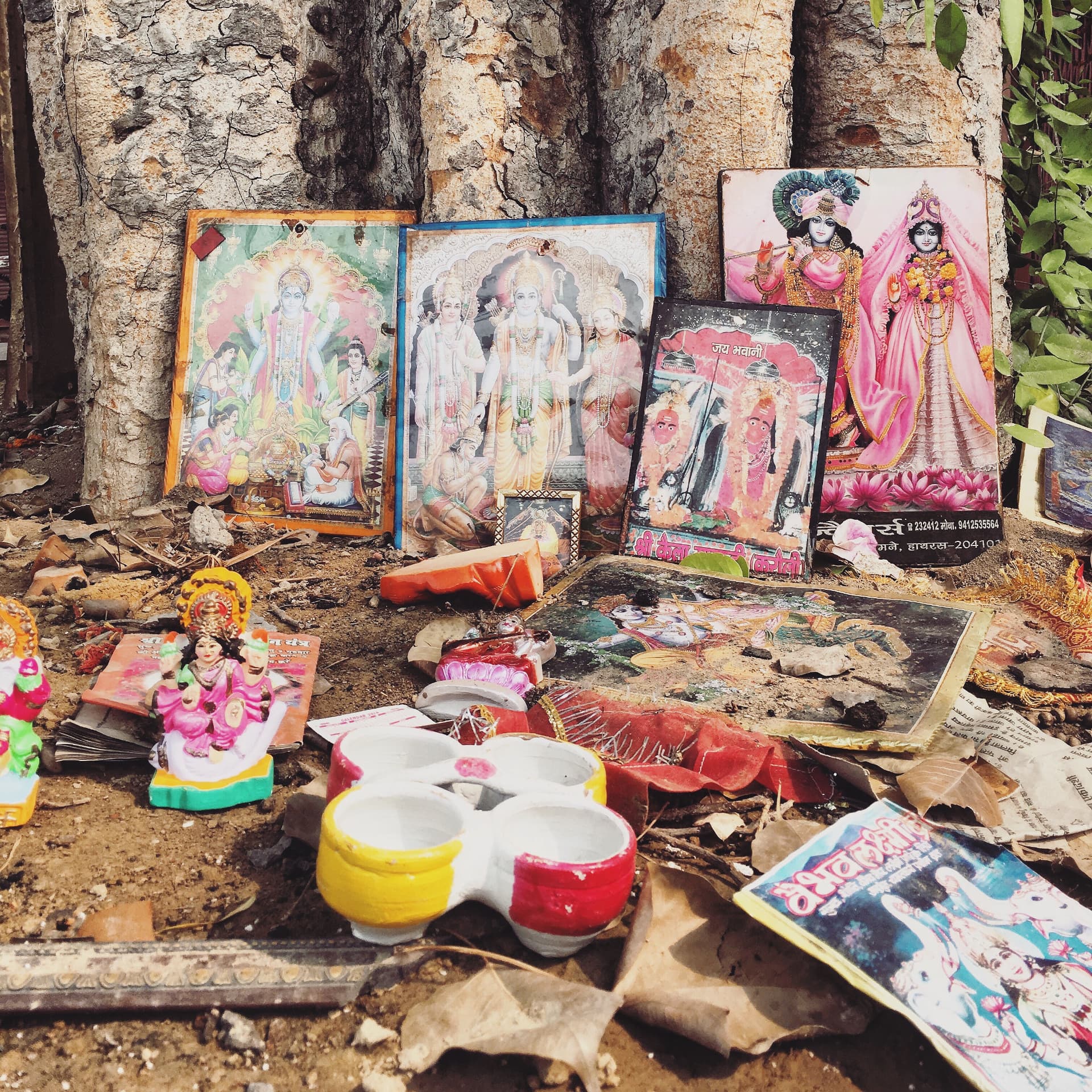A small outdoor altar at the base of a tree, with framed pictures of Hindu deities, a painted clay lamp dish and ritual booklets