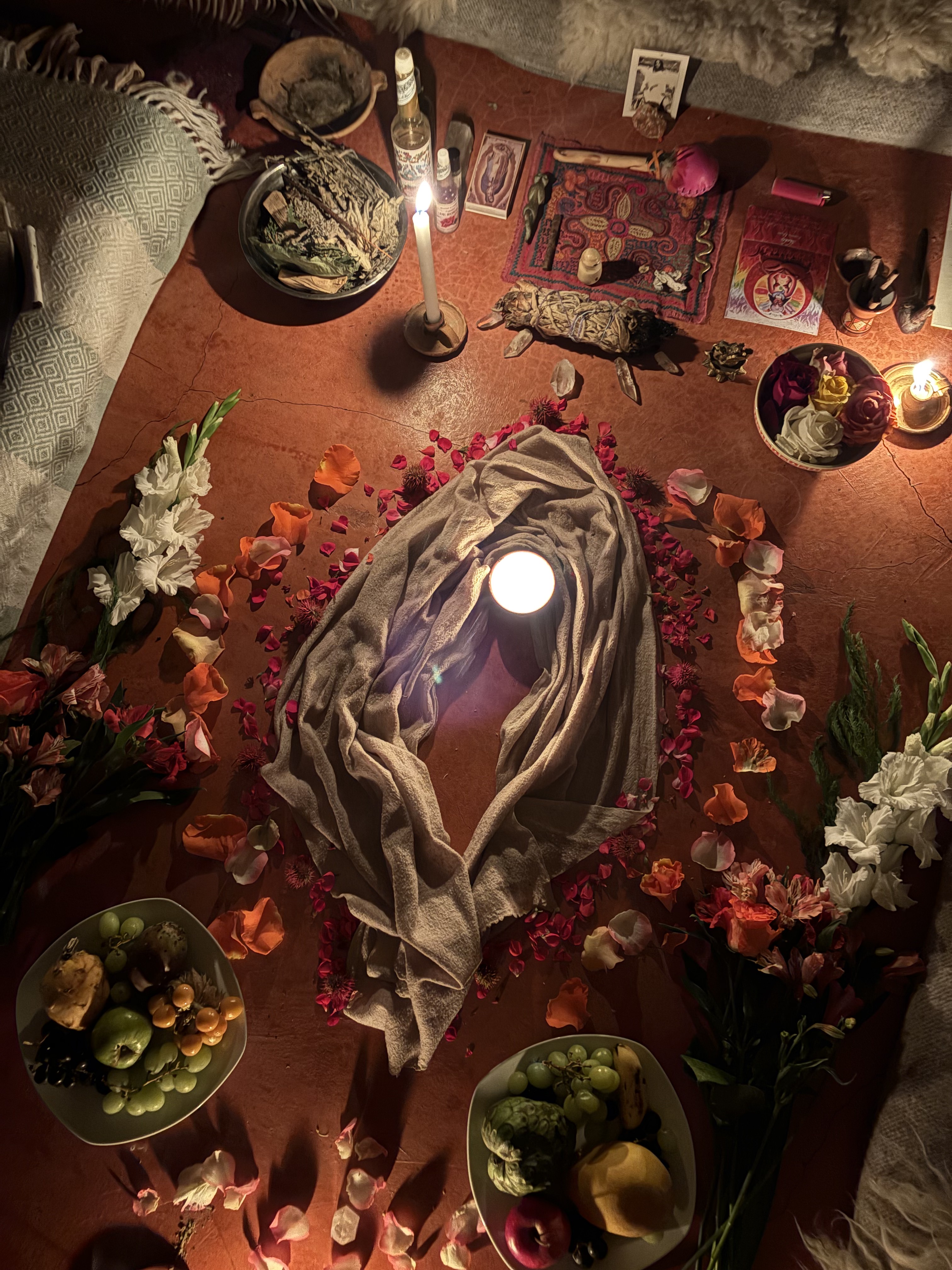 Erika's floor altar: a draped cloth cradling a lit candle, ringed with petals, flowers, crystals and bowls of fruit