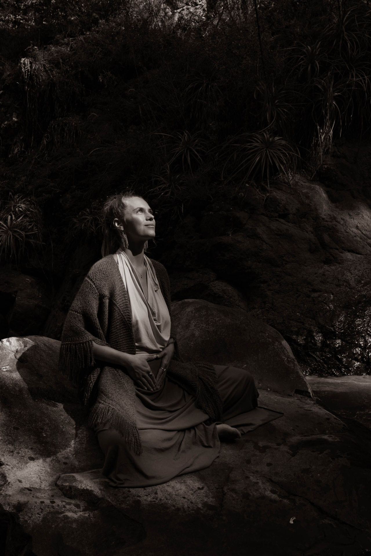 Erika Elizabeth seated on a rock with hands resting on her belly, face lifted toward the light