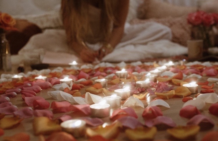Lit tea lights and scattered rose petals arranged in a spiral on the floor, with Erika seated cross-legged behind them writing in a journal